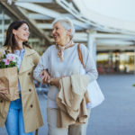 A smiling young woman helps her senior mother carry fresh groceries outside a store, exemplifying family care and support.