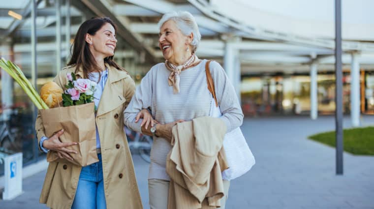 A smiling young woman helps her senior mother carry fresh groceries outside a store, exemplifying family care and support.