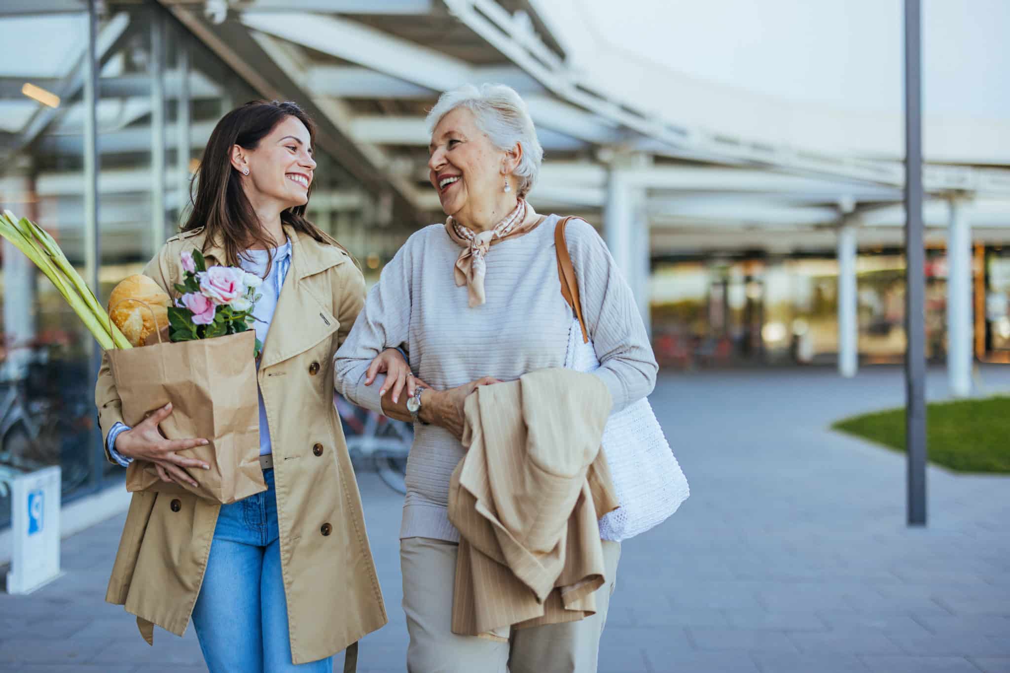 A smiling young woman helps her senior mother carry fresh groceries outside a store, exemplifying family care and support.