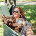 Woman reading book relaxing in hammock with her fluffy brown Maltipoo dog on sunny day