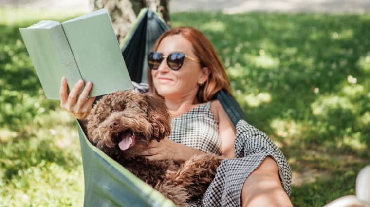 Woman reading book relaxing in hammock with her fluffy brown Maltipoo dog on sunny day