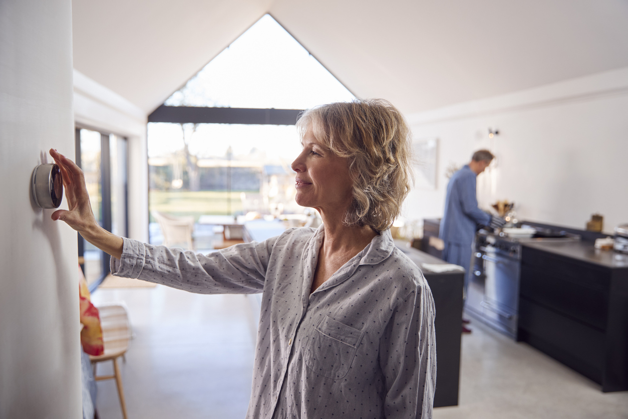 Woman adjusting a wall thermostat in a bright open-plan kitchen while a man cooks at the stove in the background.