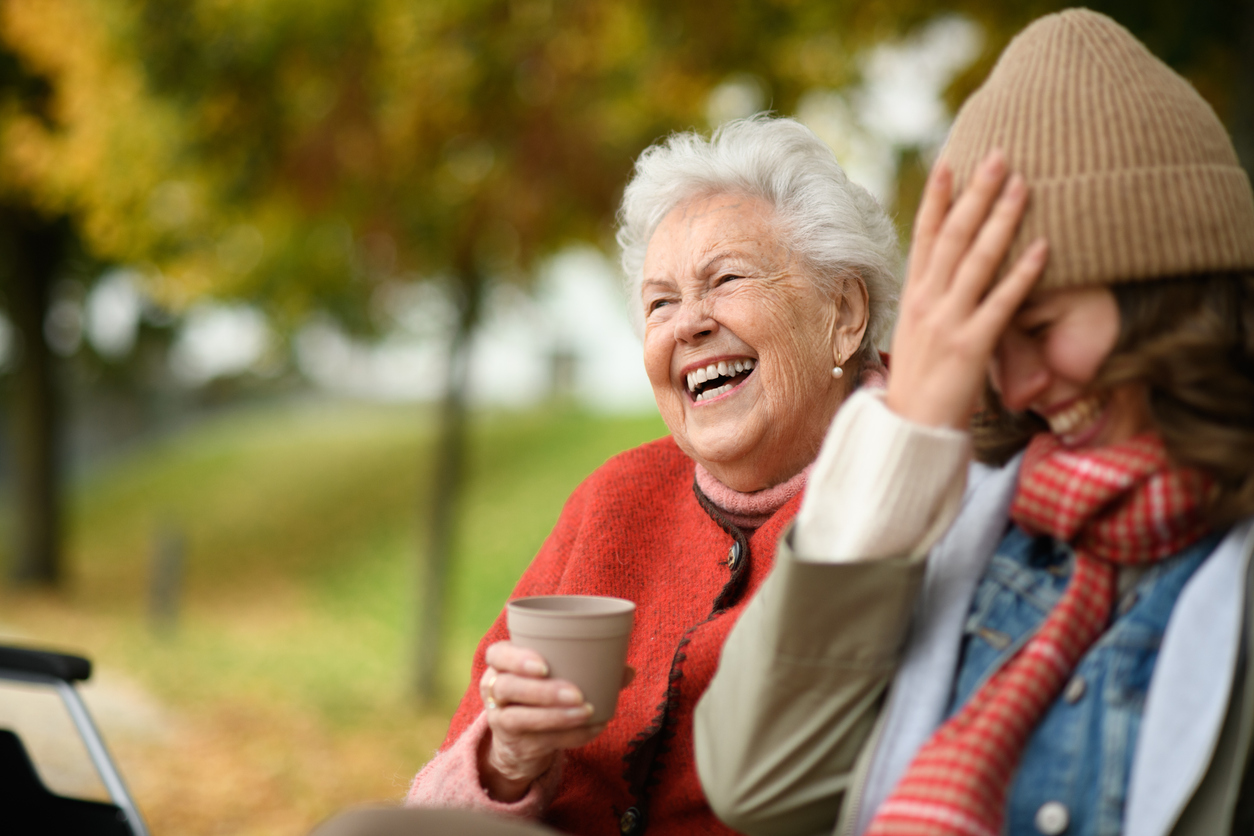 Two women laugh together outdoors on a park bench; an elderly woman with white hair holds a coffee cup.