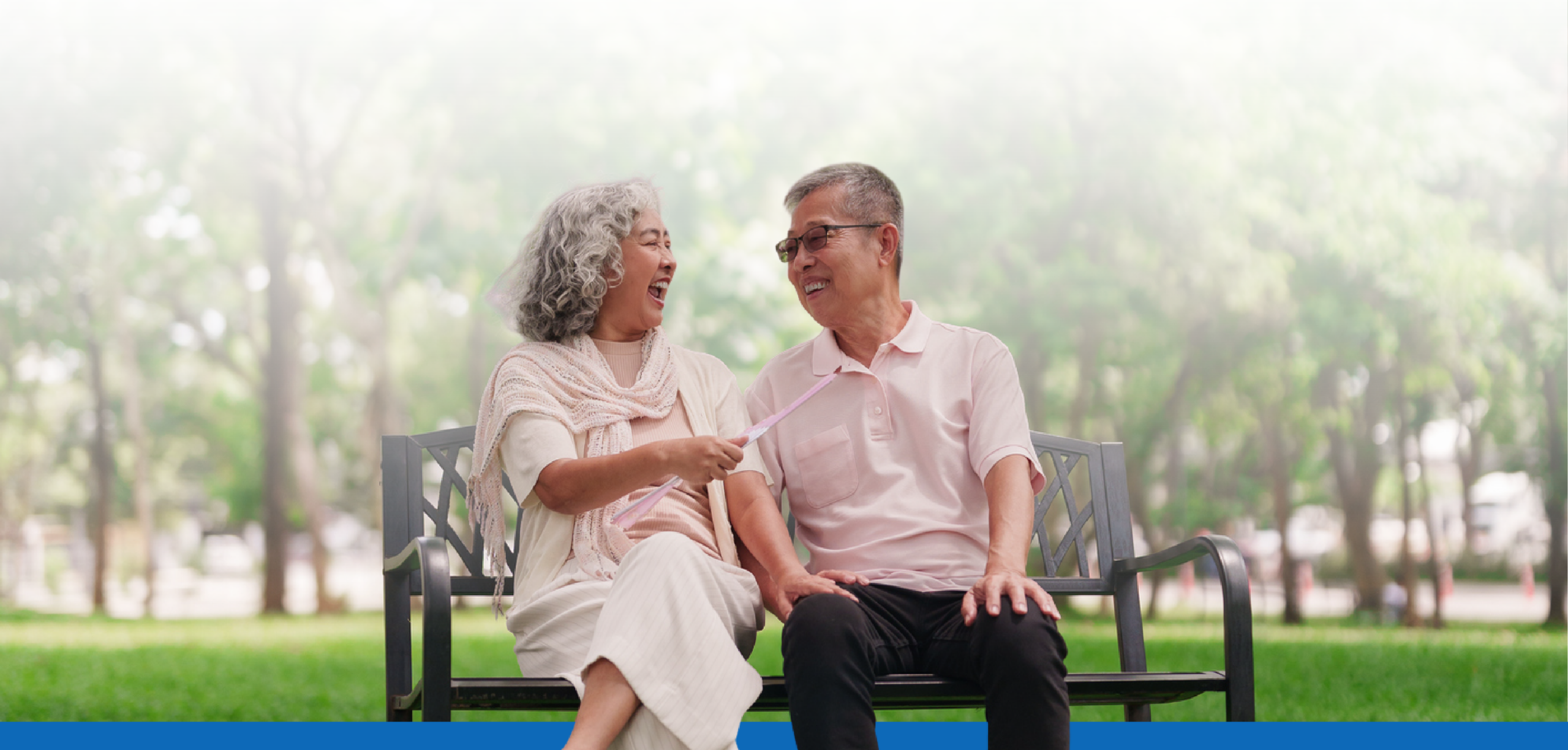 Elderly couple sitting on a park bench, smiling and laughing together outdoors.