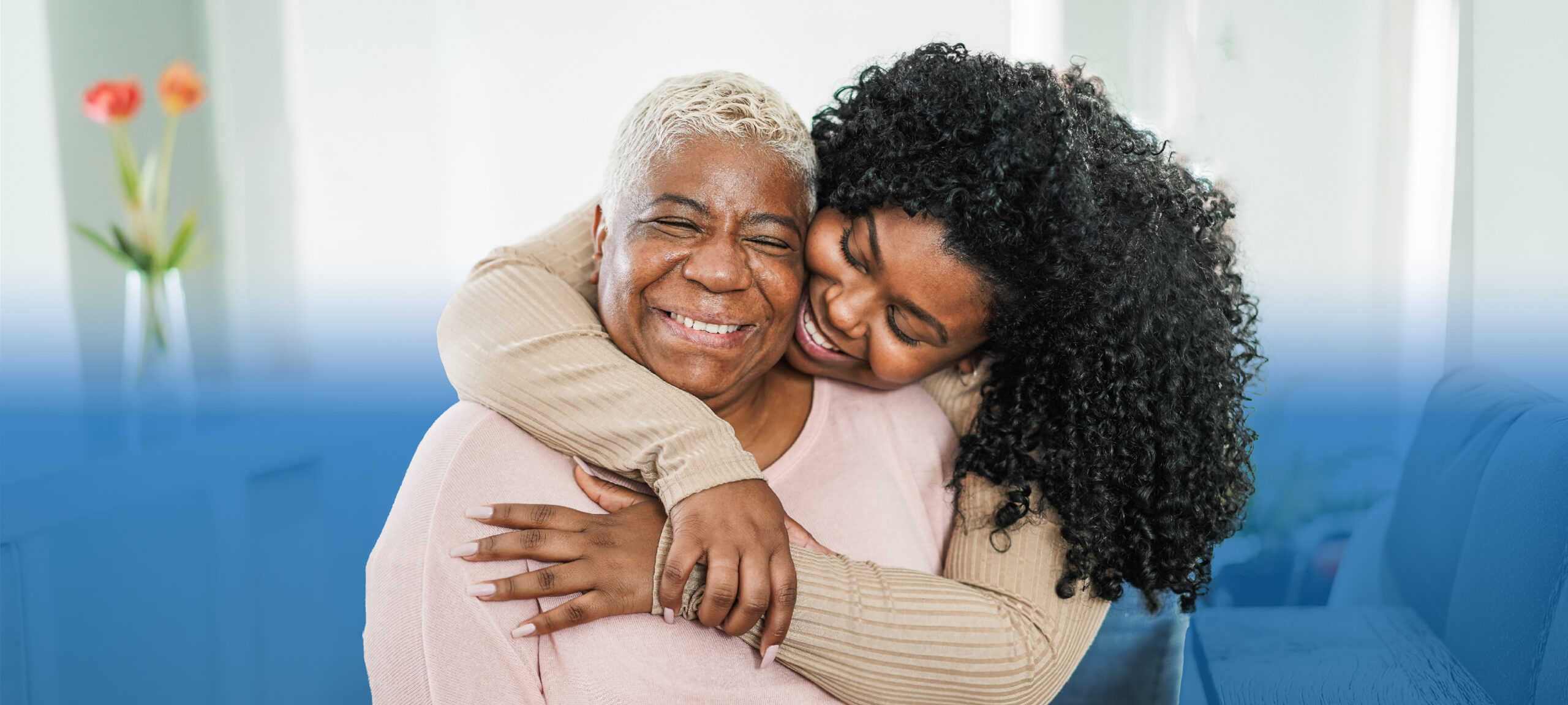 Two women hugging warmly; an older woman with gray hair smiles as a younger woman embraces her from behind.