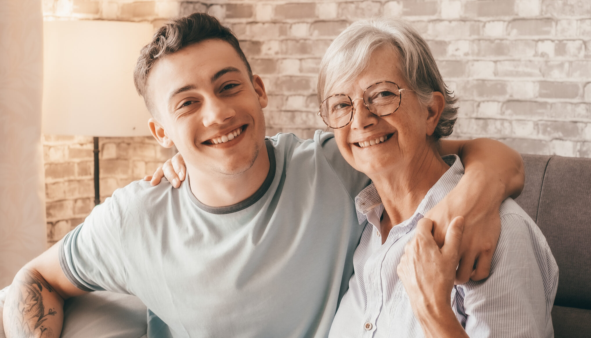 Young man and older woman smile while hugging on a sofa in a cozy living room with a brick wall backdrop