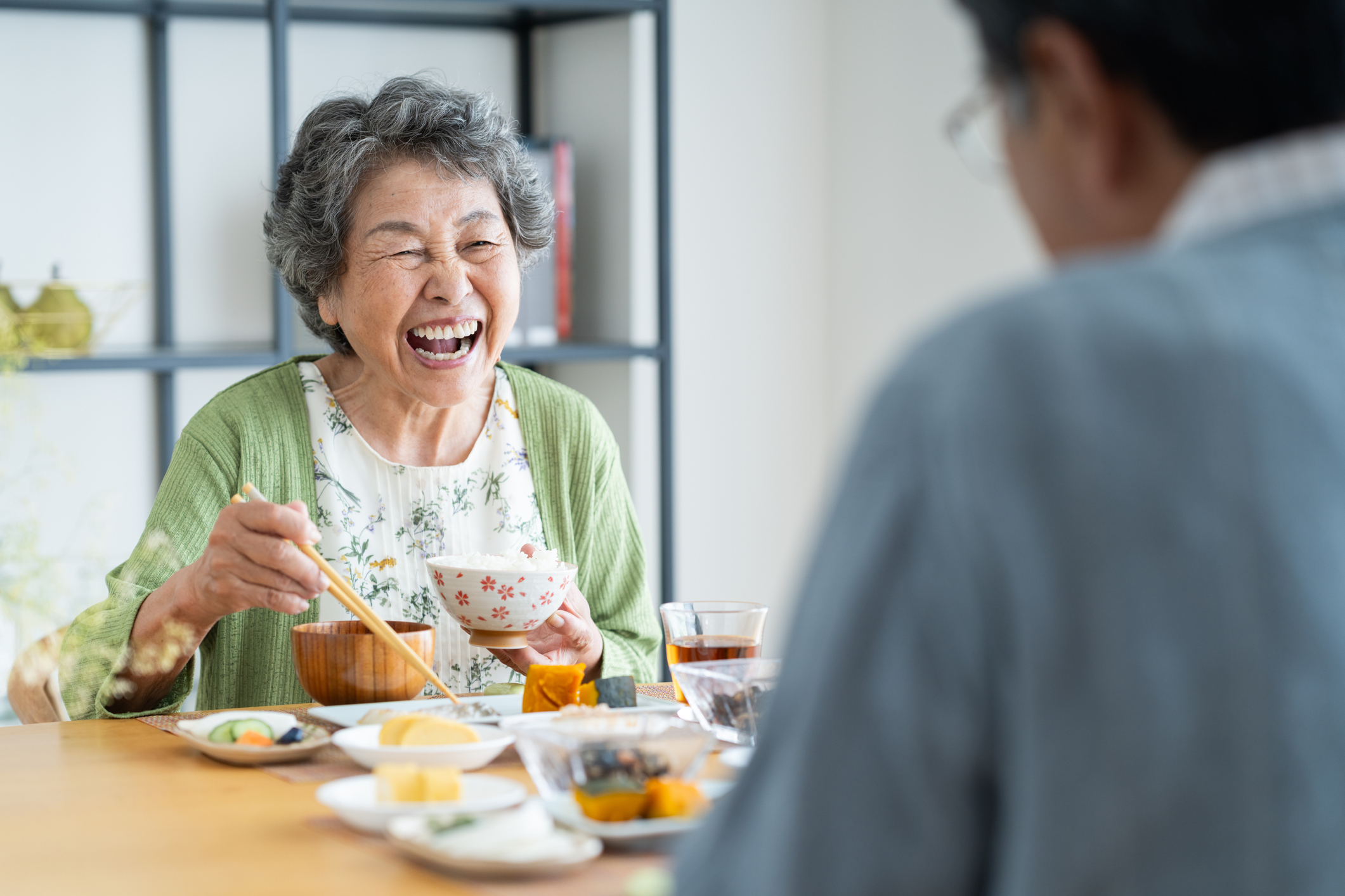 Elderly woman laughing while using chopsticks to eat at a dining table with assorted dishes; another person is blurred in the foreground.