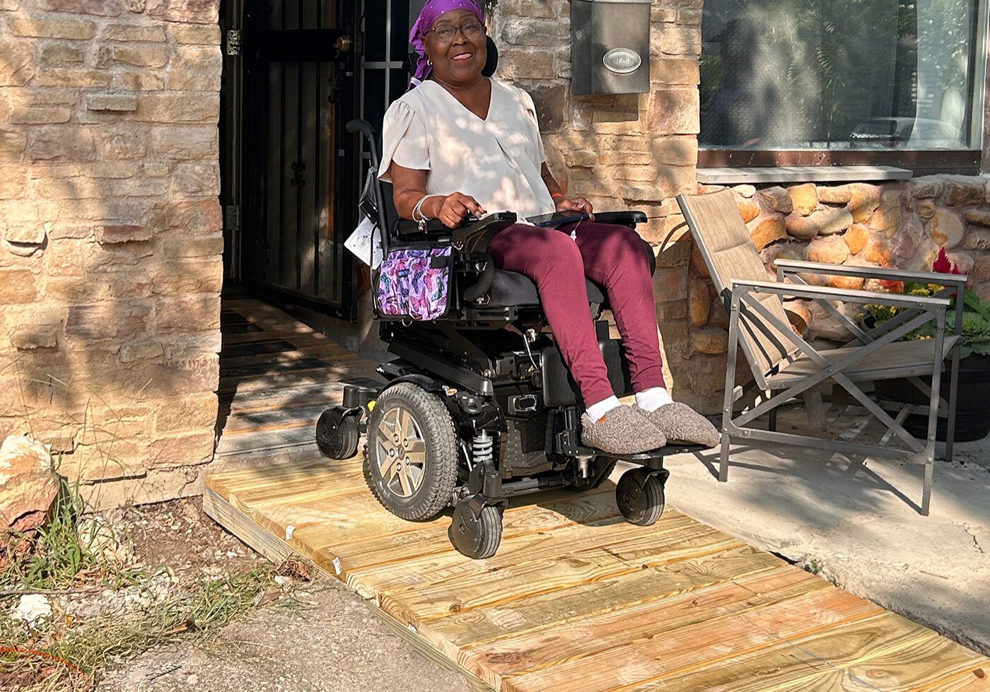 Older woman in a wheelchair on a wooden ramp outside a stone house, smiling at the camera.