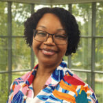 Smiling woman with short curly hair and glasses, wearing a bright multicolor patterned blouse, standing indoors by large windows.