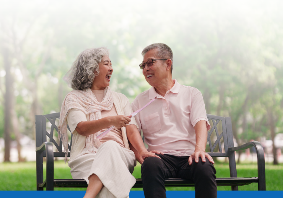 Elderly couple sitting on a park bench, smiling and laughing together outdoors.