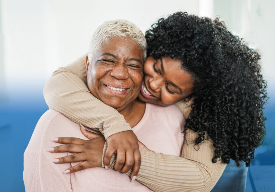Two women hugging warmly; an older woman with gray hair smiles as a younger woman embraces her from behind.