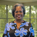 Portrait of a smiling Black woman with short gray-streaked hair wearing a blue floral blouse indoors.