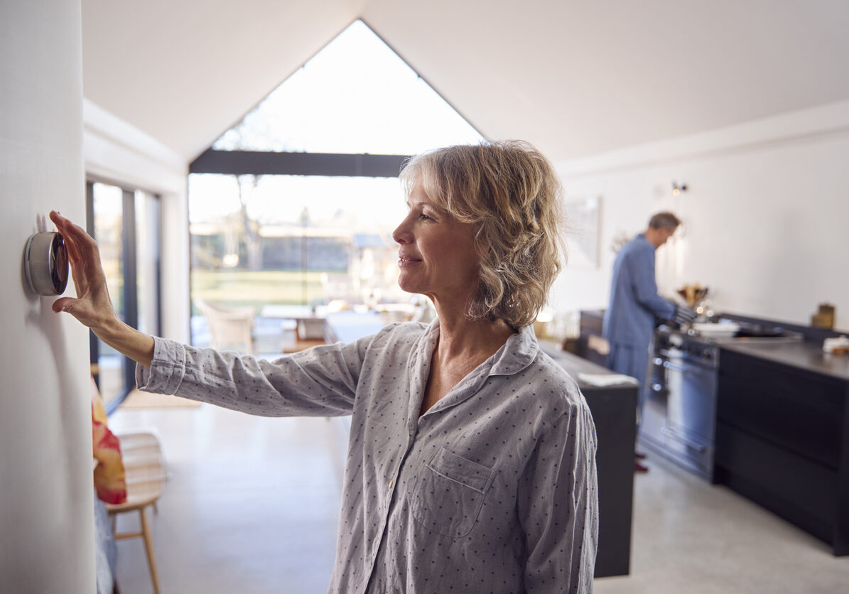 Woman adjusting a wall thermostat in a bright open-plan kitchen while a man cooks at the stove in the background.