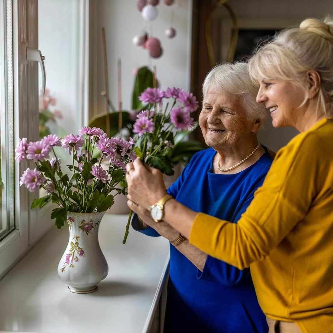 Daughter caregiver and older mother putting purple flowers in a vase