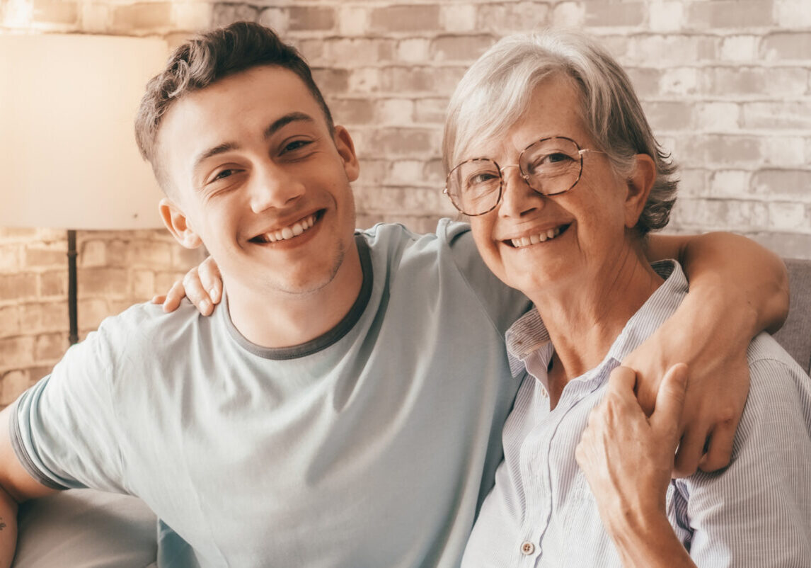 Young man and older woman smile while hugging on a sofa in a cozy living room with a brick wall backdrop