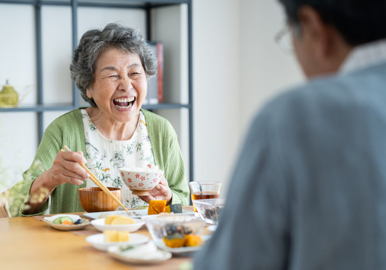 Elderly woman laughing while using chopsticks to eat at a dining table with assorted dishes; another person is blurred in the foreground.